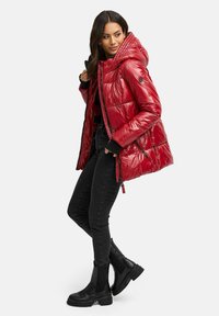 Rote Pufferjacke mit glänzender Oberfläche, Kapuze, Reißverschluss, seitlichen Taschen und gestepptem Design. Kombiniert mit schwarzen Jeans und Stiefeletten.