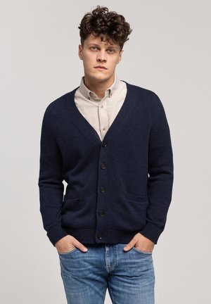 Granatowy kardigan wykonany z teksturowanej tkaniny, z dekoltem w serek, przednimi kieszeniami i ciemnymi guzikami, noszony na jasnej koszuli z guzikami.