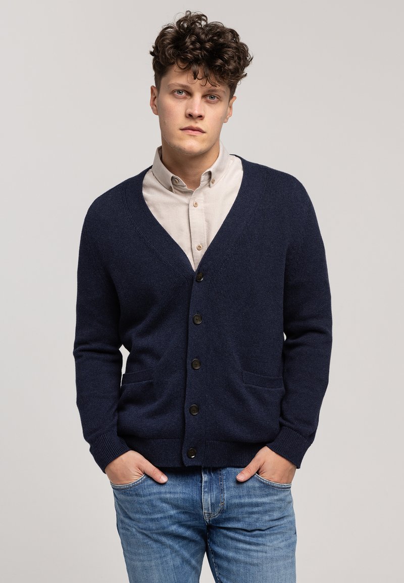 Granatowy kardigan wykonany z teksturowanej tkaniny, z dekoltem w serek, przednimi kieszeniami i ciemnymi guzikami, noszony na jasnej koszuli z guzikami.