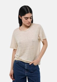 T-shirt beige à manches courtes avec un col rond, présentant une texture légèrement transparente. Associé à un jean en denim bleu foncé.