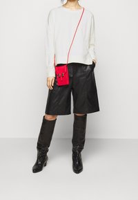 Bílá mikina, černé kožené šortky, černé kozačky po kolena a malá červená kabelka přes rameno s kovovými doplňky.