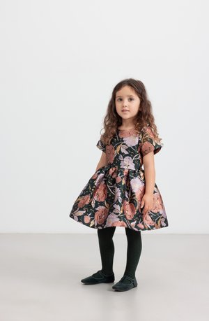 Robe florale avec un fond noir, ornée de fleurs roses et violettes, à manches courtes, avec une jupe évasée, associée à des collants verts et des chaussures noires.