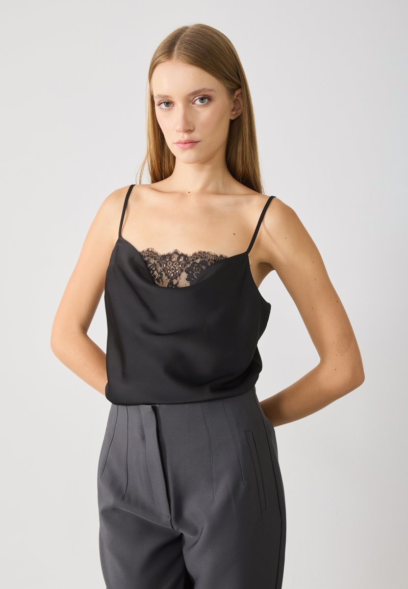 Zwarte satijnen camisole met een kanten detail langs de halslijn, dunne bandjes, gecombineerd met donkergrijze geplooide broek. Over het geheel een soepele textuur.