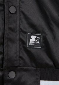 Veste en tissu noir avec un écusson rectangulaire noir et blanc portant le texte "STARTER BLACK LABEL" et des boutons-pression ronds noirs.
