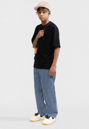 Dospievajúci nosiaci béžovú čiapku, čierne oversized tričko, modré rifle, biele tenisky, držiaci popruh kabelky cez rameno.