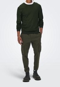 Pull vert à col rond en tissu tricoté, associé à un pantalon cargo olive avec poches latérales, et des bottes noires à lacets.