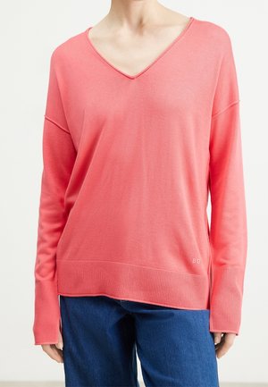 Vrouw draagt een losse koraalkleurige V-hals trui met verlengde geribde manchetten en blauwe spijkerbroek, staand tegen een effen achtergrond.