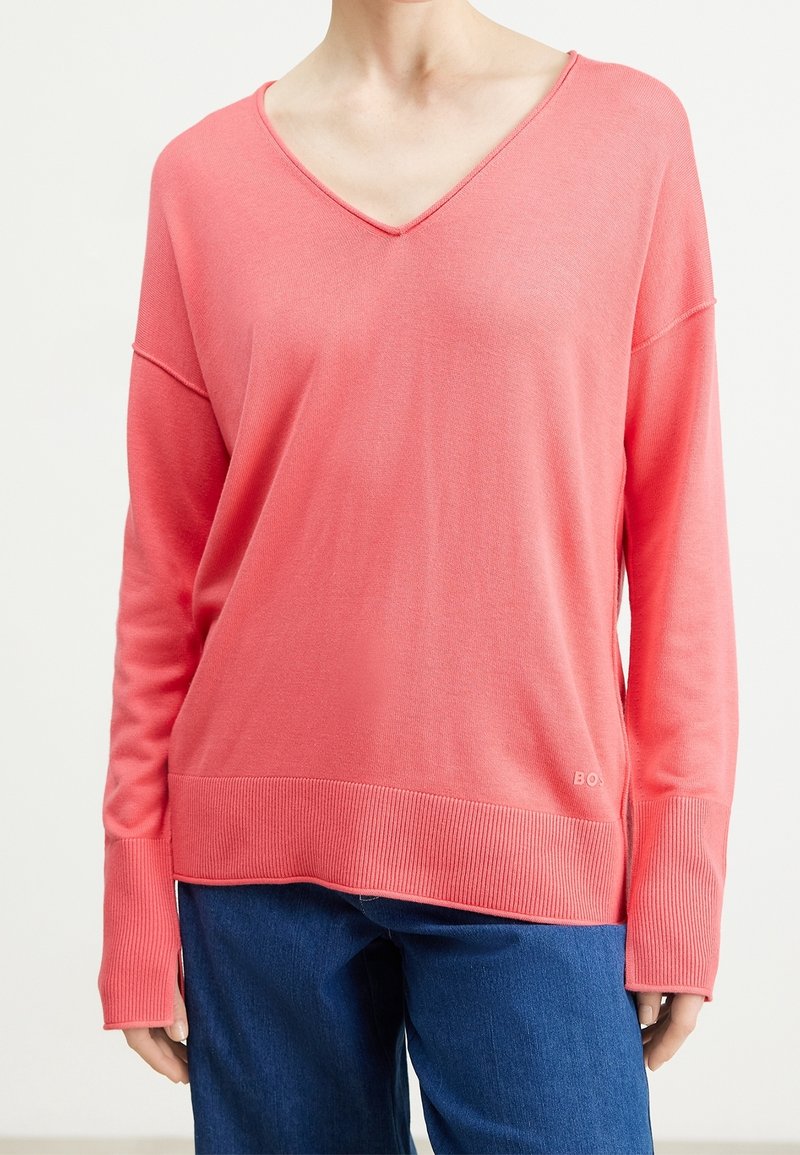 Femme portant un pull ample corail à col en V avec des poignets côtelés allongés et un jean bleu, debout devant un fond uni.