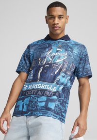 Sininen graafinen polo-paita, jossa on tummansininen kaulus, ja jossa on yksityiskohtainen painatus Marseille-logoista ja teksteistä, teksturoitua kangasta ja lyhyet hihat.