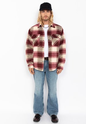 Rood en crèmekleurig geruit flanellen shirt met voorzakken, gedragen over een wit T-shirt, gecombineerd met lichtblauwe uitlopende spijkerbroek. Zwarte pet.