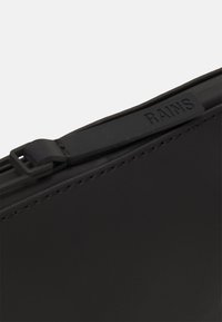 Schwarze wasserdichte Tasche mit glatter Textur, Reißverschluss und einem auffälligen, geprägten Markenetikett "RAINS". Mit verstärkten Nähten.