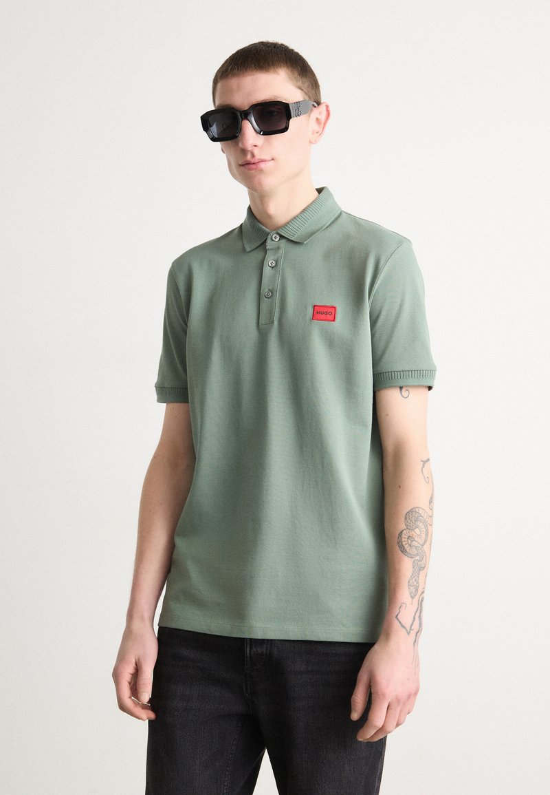 Polo-shirt i lys grønn, kortermet, med tre-knappers placket, rød logo-lapp. Brukt med svarte rektangulære solbriller. Mørke jeans.