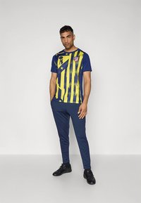 Chemise de sport bleue et jaune à rayures verticales avec logo. Associée à un pantalon bleu marine et des baskets noires. Texture lisse et coupe ajustée.