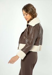 Giacca corta in pelle marrone con colletto e polsini in morbido color crema. Presenta una tasca con zip e una texture liscia.