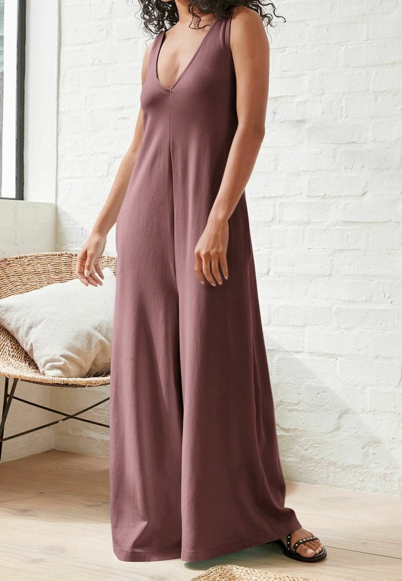 Femme portant une combinaison mauve sans manches à jambes larges, debout à l'intérieur près d'une chaise en osier avec coussin sur un sol en bois.