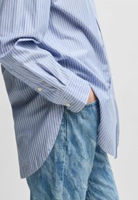 Blå och vit randig långärmad skjorta med avslappnad passform, knäppta manschetter och sidokilar, i kombination med blekta denimjeans.