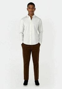 Ung mann står med hendene i lommene, iført en hvit skjorte med knapper, brune bukser og svarte sneakers mot en enkel, lys bakgrunn.