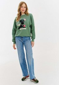 Zielona bluza z grafiką psa i kwiatowymi akcentami, w połączeniu z jasnoniebieskimi dżinsami oraz zielono-czarnymi trampkami.