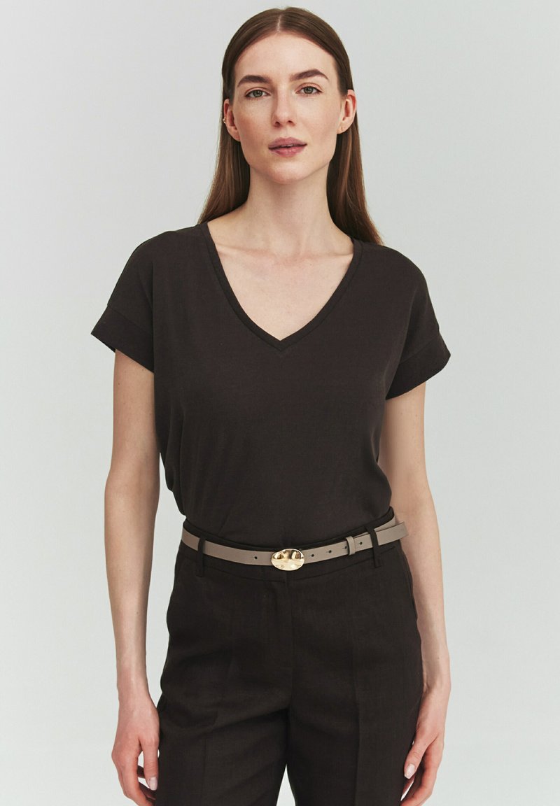 Femme aux longs cheveux bruns portant un t-shirt noir à col en V, un pantalon noir et une ceinture beige avec une boucle ovale dorée.