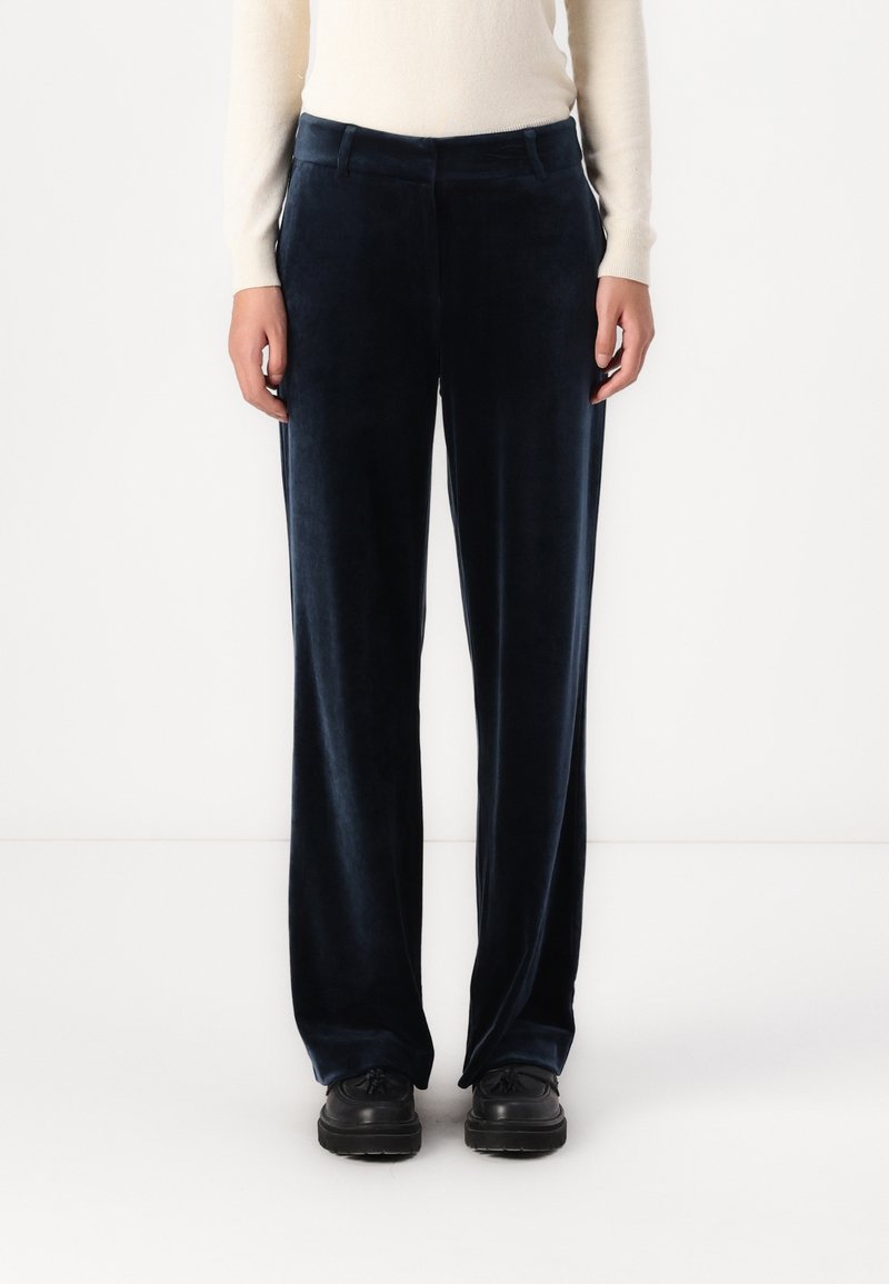Personne portant un pantalon large en velours bleu marine foncé, des chaussures noires et un haut à manches longues beige clair, debout devant un fond blanc uni.