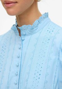 Svetlo modra bluzica z valovitim ovratnikom, gumbi na sprednjem delu ter zapletenim vezenjem s izrezi in teksturiranimi vertikalnimi črtami.