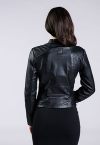 Giacca di pelle nera con design aderente, leggera lucentezza e un piccolo dettaglio con logo sul retro. Texture liscia e silhouette su misura.