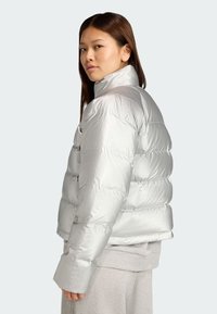 Stříbrná puffer bunda s lesklou prošívanou texturou, vysokým límcem a širokými manžetami. Nosená přes šedý svetr, ukazuje uvolněný střih.