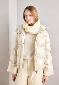 Béžová péřová bunda s lesklým povrchem, předním zipem a patentkami. V kombinaci s měkkým, off-white, žebrovaným pleteným šátkem a světlým topem.