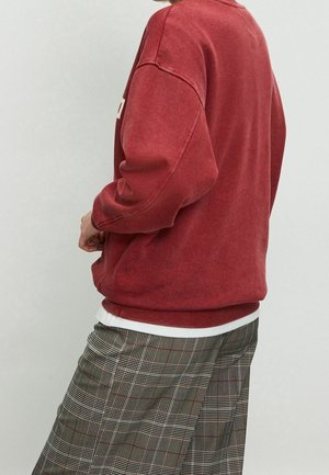 Red cotton sweatshirt with ribbed cuffs and hem, worn over a white shirt, paired with plaid trousers featuring thin lines of red and black.