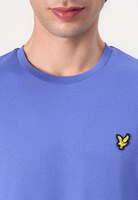T-shirt en coton violet avec un col rond et des manches courtes. Présente un logo d'aigle brodé jaune sur le côté gauche de la poitrine.