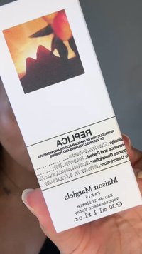 Hand die Maison Margiela Replica parfumdoosje vasthoudt met een bloemige zonsonderganglabel en geuromschrijving in zwarte tekst op een witte achtergrond.