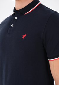 Polo blu navy con colletto a righe bianche e rosse, chiusura a tre bottoni e piccolo emblema rosso a forma di uccello sul petto.