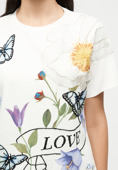 Λευκό πουκάμισο με κεντητά μεγάλα λευκά και μοβ λουλούδια, μπλε πεταλούδες και μαύρο κείμενο «LOVE» με χάντρες μπροστά.