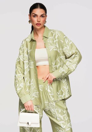 Femme portant une tenue texturée florale vert clair avec chemise ample et pantalon, top court blanc, tenant un petit sac à main blanc sur fond uni.