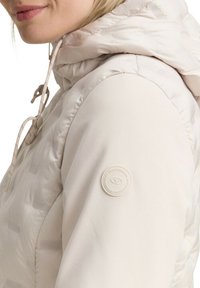 Veste matelassée beige clair avec capuche, présentant une finition texturée, des cordons de serrage et un patch logo circulaire sur la manche.