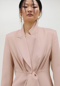 Svijetlo ružičasti sako krojen s detaljem čvora na struku, s oštrim reverima i teksturiranim materijalom. Minimalistički dizajn s čistim linijama.