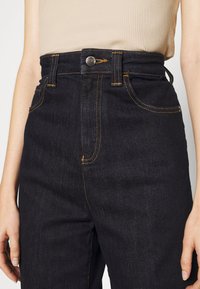 Högmidjade mörka jeans i denimtyg med synlig gul sömnad, femficka-design och en silvrig knappstängning i midjan.