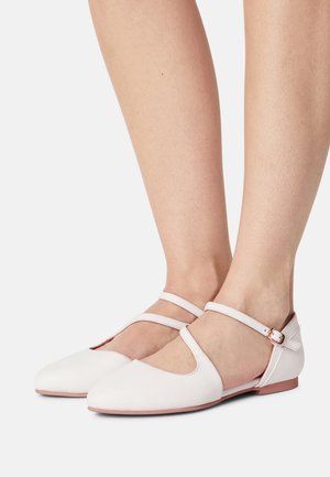 Pieds portant des chaussures plates blanches avec doubles brides et boucles en or rose, sur un fond blanc.