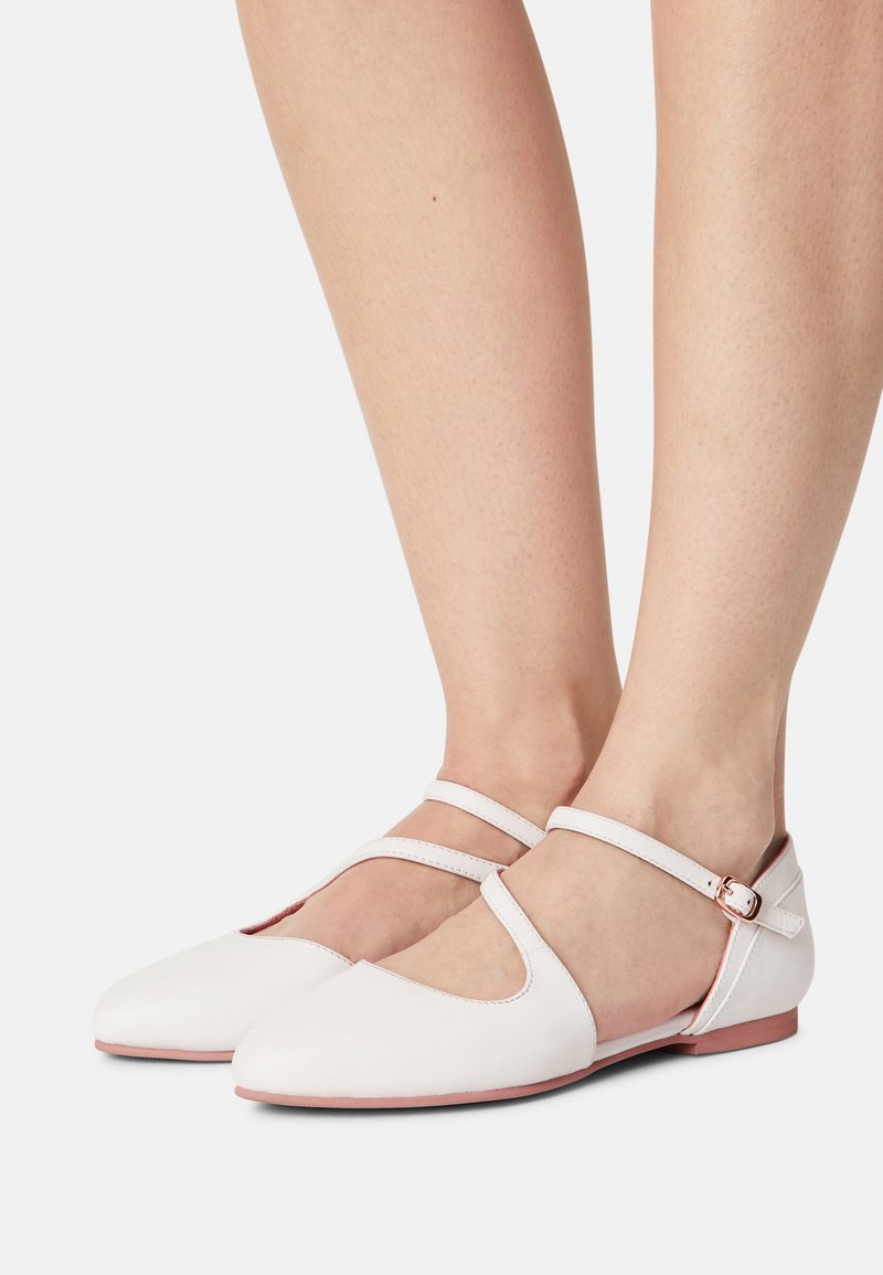 Pieds portant des chaussures plates blanches avec doubles brides et boucles en or rose, sur un fond blanc.