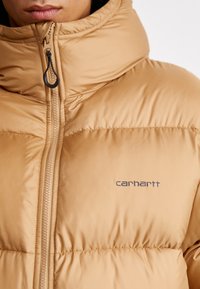 Tan pufferjakke med høj krave, lynlås foran og synlige sømme. Har et Carhartt-logo på venstre side. Glat, polstret tekstur.