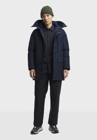 Marineblauwe gewatteerde jas met een grote capuchon, rits aan de voorkant en zijzakken, gecombineerd met een zwart shirt, zwarte broek en grijs-zwarte sneakers.