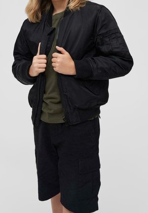 Chaqueta bomber negra con puños acanalados, cremallera frontal y bolsillos laterales, llevada sobre una camiseta verde oliva y pantalones cortos negros. Textura mate en toda la prenda.