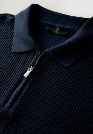 Marinblå ribbad polo med dragkedja framtill, med en slät krage och dragkedja i silver. Mjuk textur och diskret design.