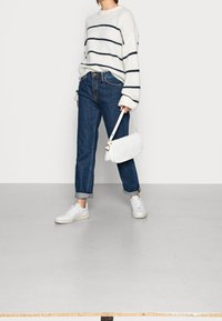 Pull en maille blanche à rayures bleu marine, jean en denim bleu et baskets blanches. Le modèle porte un sac à main matelassé blanc, debout sur un sol clair.