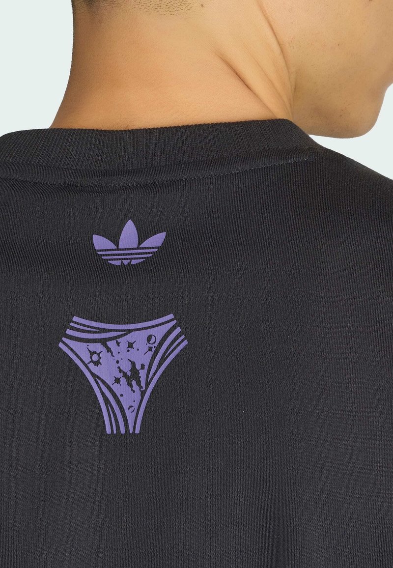 Person trägt ein schwarzes Shirt mit einem lila Adidas-Trefoil-Logo und einer stilisierten Unterwäsche-Grafik auf dem oberen Rücken.