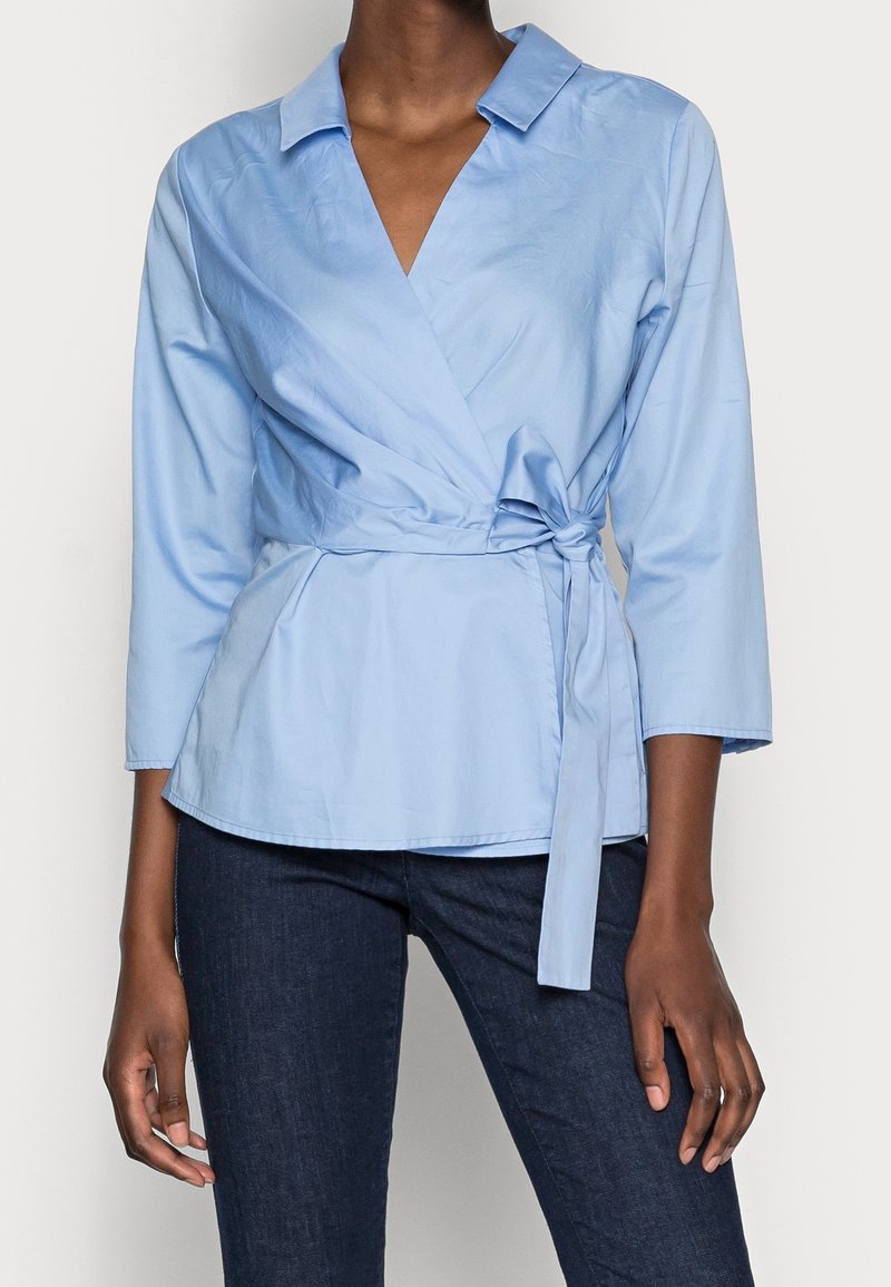 Femme portant un chemisier cache-cœur bleu clair à manches trois quarts et un jean bleu foncé sur fond blanc.
