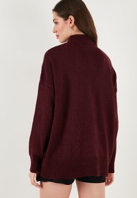 Burgundi színű kötött pulóver magas nyakkal, ejtett vállakkal és bordás aljjal. Puha tapintású és laza szabású. Hátulnézet látható.