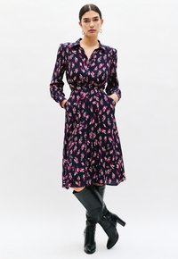 Robe à manches longues, de couleur marine avec un motif floral rose, taille ceinturée et poches latérales. Associée à des bottes noires jusqu'aux genoux. Tissu doux, design avec col.