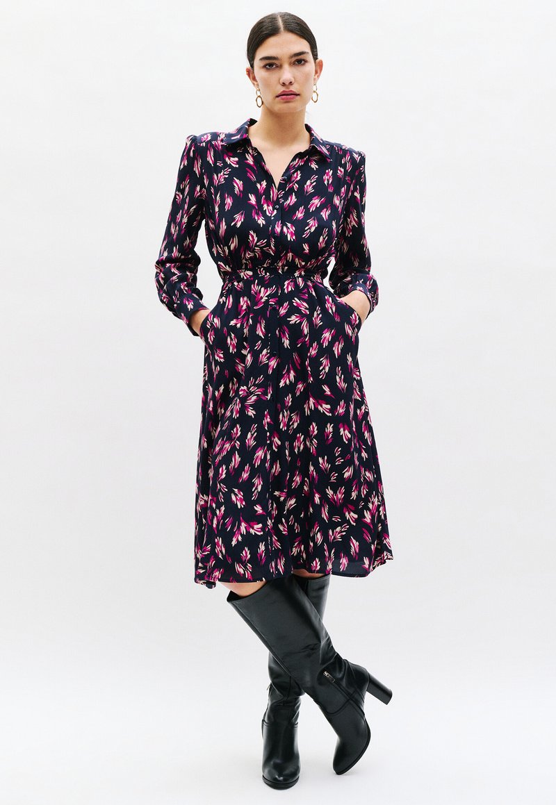 Robe à manches longues, de couleur marine avec un motif floral rose, taille ceinturée et poches latérales. Associée à des bottes noires jusqu'aux genoux. Tissu doux, design avec col.