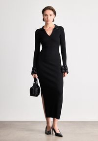 Vestido preto canelado com decote em V, colarinho ornamentado e mangas sino; apresenta uma fenda até a coxa e é combinado com uma pequena bolsa preta.